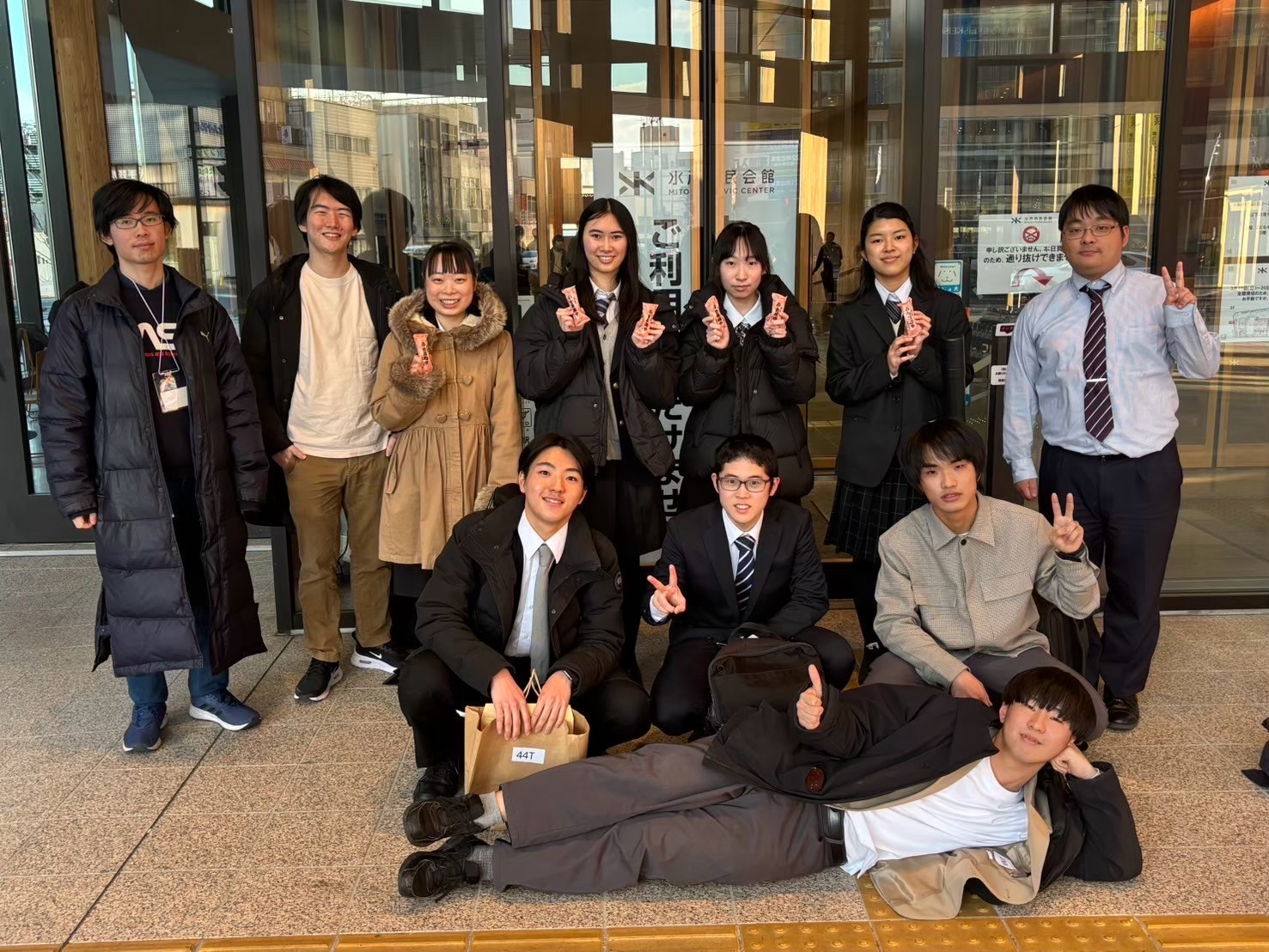 会場前で記念撮影をするA班の生徒と班長・TA。2列に並んで写っているが、前で横になる受講者がいる。TA の一人は若干場違いなスーツを着ている。